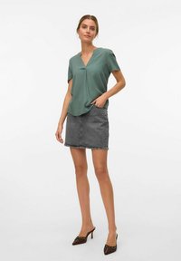 Blouse verte à manches courtes avec un décolleté en V, associée à une jupe en denim noire avec des accents cloutés et des mules à talons hauts marron.