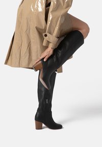 Stivale in pelle nera fino alla coscia con finitura testurizzata, zip laterale e tacco a blocco in tonalità legno naturale. È visibile parte di un trench coat beige lucido.