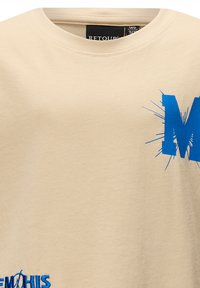 Beżowa bawełniana koszulka z okrągłym dekoltem, z dużym niebieskim nadrukiem litery "M" i wybuchowymi akcentami, o miękkiej strukturze.