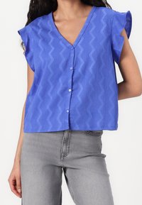 Modrá košile na knoflíky s krátkým rukávem a výstřihem do V, se strukturovaným vzorem ve tvaru cik-cak a pohodlným střihem. V kombinaci se šedými džíny.