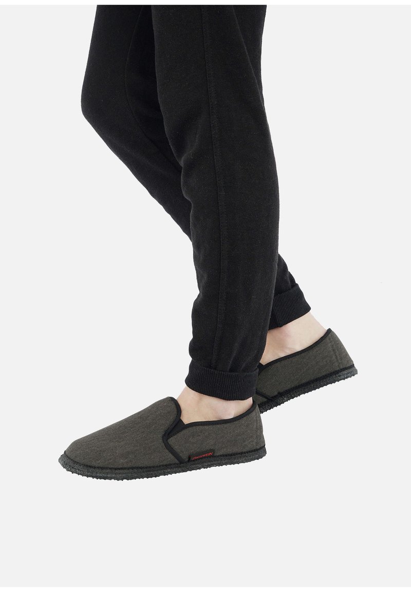 Baskets en tissu gris à enfiler avec des accents noirs et des semelles en caoutchouc texturé, portées avec un pantalon de jogging noir. Design minimaliste, sans lacets visibles.