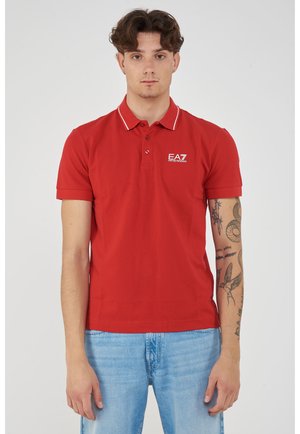 Uomo con capelli corti e ricci, indossa una polo rossa EA7 e jeans blu, in piedi con il braccio destro tatuato visibile su sfondo bianco.