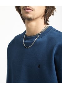 Donkerblauwe sweatshirt met een geribbelde ronde hals en een subtiel zwart logo. Beschikt over een gelaagde look met een grijze onderhemden en een zilveren ketting.
