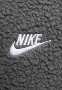 Pelēka vilnas auduma virsma ar tekstūru; tai ir izteikta balta izšūta Nike logo, kas novietots labajā pusē.
