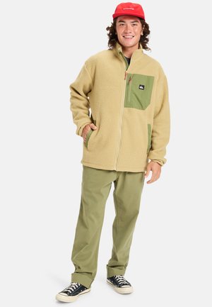 Fleece jacket in beige with green pocket accents. Set against olive green pants and black sneakers. Zip closure and side pockets visible.