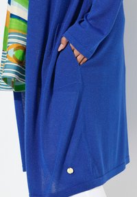 Blauwe cardigan van een zachte, gestructureerde stof, met een zak en een gouden accent detail bij de zoom. Gelaagd met een geblokte top.