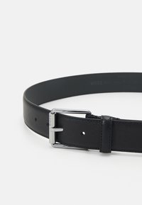 Ceinture en cuir noir avec une surface texturée, dotée d'une boucle en métal argenté et d'un branding subtil. Comprend des trous d'ajustement pour la taille.