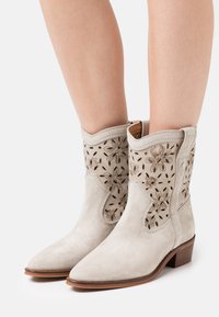 Light gray suede ankle boots with floral cut-out design, pointed toe, and low wooden heel. Features stitched accents along the edges.