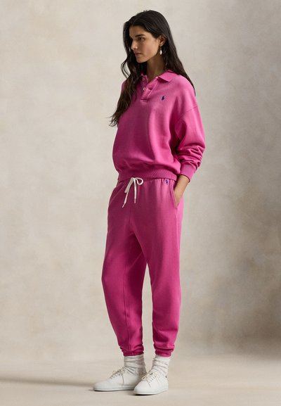 Ροζ βαμβακερό φούτερ με γιακά και λογότυπο, σετ με αντίστοιχο παντελόνι φόρμας με ελαστική μέση και μανσέτες στους αστραγάλους. Λευκά αθλητικά παπούτσια.