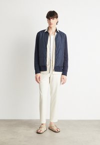 Chaqueta bomber navy con textura acolchada, cuello crema, camisa blanca debajo, pantalones de color claro y sandalias marrones con tiras.