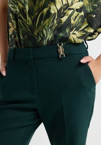 Groene op maat gemaakte broek met een gladde textuur, zijzakken en een unieke gouden accentgesp op de tailleband. Bloemige blouse gedeeltelijk zichtbaar.