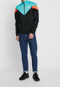 Chaqueta negra con diseño de bloques de color aqua y naranja, cierre de cremallera y puños acanalados, combinada con vaqueros azules y zapatillas negras.