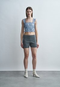 Νεαρή γυναίκα που φοράει μπλε καρό κομμένο τοπ, γκρι σόρτς με υφή και μικρές κουκκίδες, λευκές κάλτσες και λευκά παπούτσια με τακούνι, στέκεται σε γκρι δάπεδο.