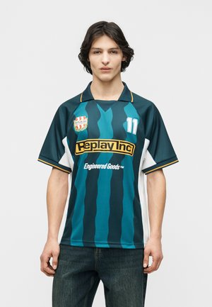 Giovane che indossa una maglia sportiva a righe blu-verde con il logo "Replay Inc" e il numero 11, in piedi contro uno sfondo semplice.