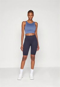 Kék bordázott rövid felső, magas derekú sötét leggingssel párosítva. A modell fehér sportzoknit és cipőt visel, egyszínű háttér előtt áll.