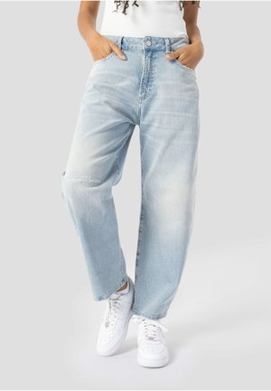 Person iført lyseblå løse jeans med hænderne i lommerne og hvide sneakers står på en lysegrå baggrund.