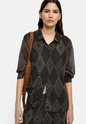 Kobieta ubrana w czarną, prześwitującą koszulę zapinaną na guziki oraz dopasowaną spódnicę z geometrycznymi wzorami diamentów, niosąca brązową torebkę na ramię z frędzlami.