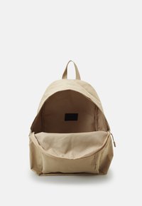 Mochila de tela beige con parte superior redondeada, tirantes ajustables y un compartimento principal que cuenta con un cierre de cremallera y un bolsillo plano en el interior.