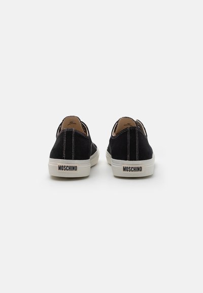 Scarpe da canvas nere con suole bianche, caratterizzate da cuciture a contrasto e un'etichetta "Moschino" sul tallone. Design semplice e a bassa altezza.