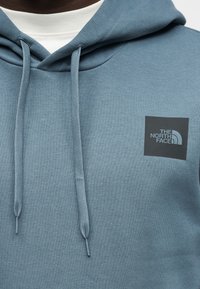 Μπλε φούτερ με κουκούλα από μαλακό ύφασμα που διαθέτει κορδόνι στην κουκούλα και μαύρη κονκάρδα με λευκό κείμενο στο στήθος.