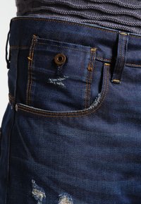 Jeans de mezclilla azul oscuro con ligero desgaste, costuras doradas y un detalle en el bolsillo frontal con un remache de metal.