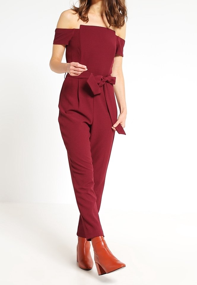 Combinaison bordeaux à épaules dénudées avec un décolleté carré, une taille ceinturée et des jambes fuselées. Associée à des bottines à talons marron.