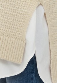 Pull en tricot couleur crème avec un design texturé, à ourlet côtelé, superposé sur une blouse blanche, associé à un jean en denim.