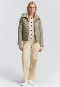 Olijfgroene pufferjack met een geborduurd patroon en capuchon. Draagt over een crèmekleurige trui en gecombineerd met beige wijde broek en witte sneakers.