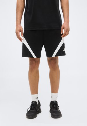 Mężczyzna ubrany w czarne sportowe spodenki z białymi ukośnymi paskami, czarne trampki, białe skarpetki Adidas oraz czarną koszulkę, stojący na białym tle.