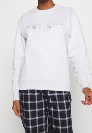 Personne portant un sweat-shirt gris clair avec le logo "carhartt" et un pantalon à carreaux noir et blanc, debout sur un fond neutre.