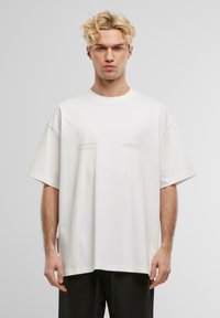 Hvit bomulls t-skjorte med oversized passform, senkede skuldre og minimal grafisk tekst på brystet. Jevn tekstur og avslappet form.