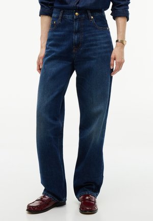 Person trägt dunkelblaue Jeans mit geradem Bein, bordeauxfarbene Loafers, ein Jeanshemd mit hochgekrempelten Ärmeln und eine goldene Armbanduhr.