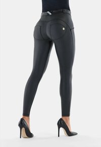 Black faux leather leggings with a form-fitting design, featuring stitching accents and two back pockets, paired with black high-heeled shoes.
