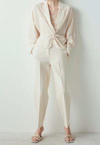 Chemise boutonnée rayée beige clair nouée à la taille, assortie à un pantalon crème fuselé et des sandales à talons blanches avec des accents dorés.