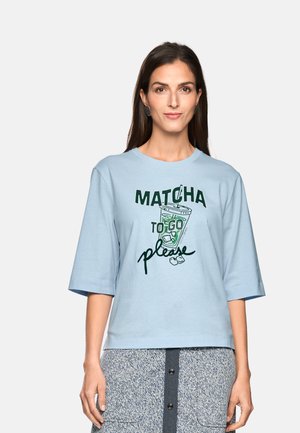 Frau trägt ein hellblaues Shirt mit dem Text "MATCHA TO GO please" und einer Grafik eines Eistees, kombiniert mit einem gemusterten Rock.