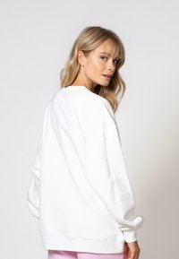 Sweat-shirt blanc surdimensionné avec épaules tombantes, poignets côtelés et une texture douce, montré de dos, avec des détails de couture subtils.