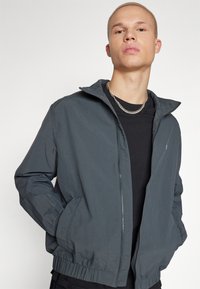 Chaqueta gris, ligera, con cierre de cremallera, dobladillo elástico y bolsillos laterales. Fabricada con una tela suave y presenta un diseño minimalista.