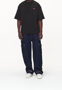 Camiseta negra de gran tamaño con un pequeño logo rojo, combinada con pantalones cargo de mezclilla azul sueltos que tienen bolsillos laterales y zapatillas blancas.