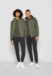 Due modelle indossano giacche leggere color verde oliva abbinate con dettagli gialli sui zipper e jogger neri, abbinate a sneakers bianche.
