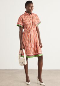 Kurzarmhemd Kleid in Orange mit Obstmotiv, grüner Borte und einem Taillengürtel. Kombiniert mit einer kleinen weißen Handtasche und weißen Absätzen.