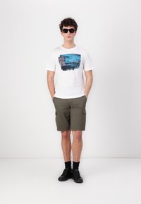 Hvit T-skjorte med blå grafisk trykk, grønne cargo-shorts, svarte joggesko og mørke solbriller, som står mot en nøytral bakgrunn.