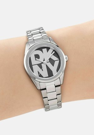 Montre-bracelet en métal argenté avec bracelet à maillons, cadran noir comportant de grandes lettres blanches DKNY qui se chevauchent, portée sur un poignet à peau claire.