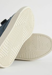 Blå texturerad sneaker med vit gummisula, med ett geometriskt mönster på sulan och remförslutning på sidan.