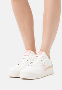 Zapatillas de cuero blanco con detalles en rosa, puntera redonda, diseño con cordones y suela texturizada. Presentan un logo en relieve en el lateral y el talón.