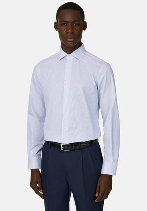 Mann trägt ein tailliertes hellblau gestreiftes Hemd mit weißen Knöpfen, kombiniert mit dunkelblauen Hosen und einem schwarzen Gürtel, steht aufrecht.