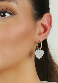 Vita hjärtformade örhängen gjorda av texturerat material, hängande från en guldkedja med en liten strassdetalj.