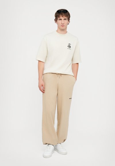 T-shirt beige avec logo sur la poitrine, associé à un pantalon beige ample avec taille élastique, et des baskets blanches. Lunettes de soleil noires sur la tête.