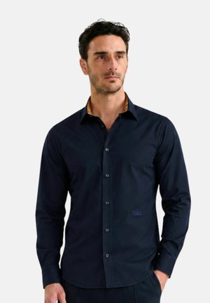 Uomo che indossa una camicia aderente blu scuro con bottoni e colletto interno a contrasto, in piedi con una mano in tasca su uno sfondo semplice.