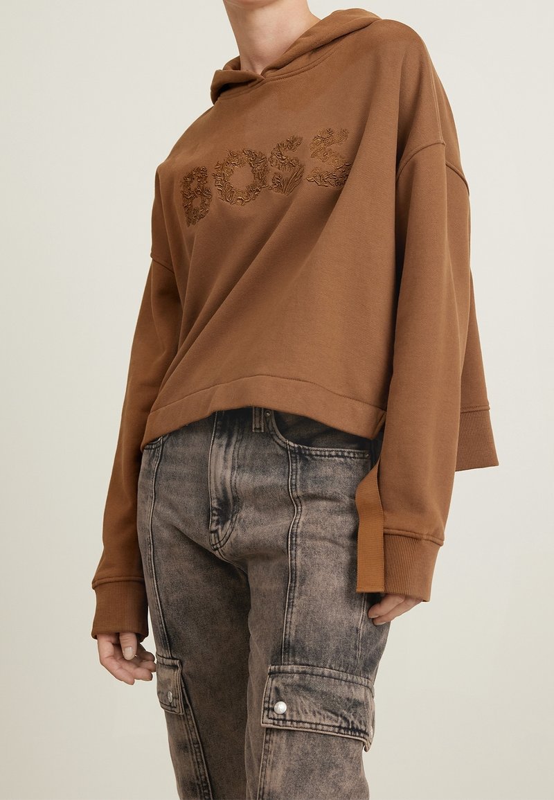 Personne portant un sweat à capuche marron avec un lettrage texturé "DOSS" et un pantalon cargo gris délavé avec des poches avant à rabat.