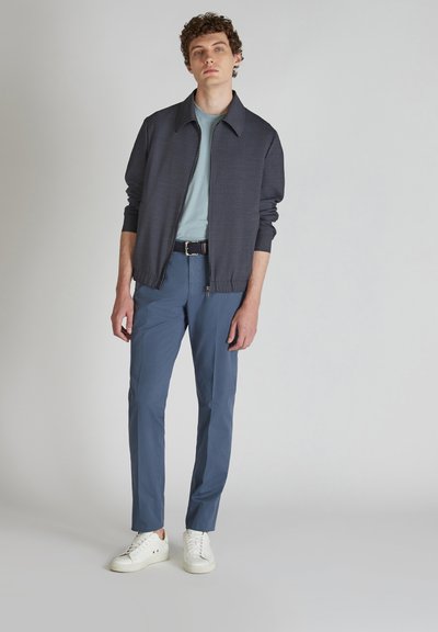 Outfit casual da uomo composto da una giacca nera con zip, t-shirt azzurro chiaro, pantaloni blu e sneakers bianche. La giacca ha un colletto.